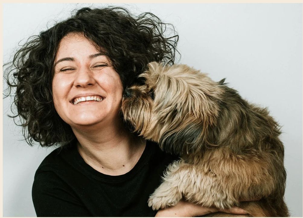 A woman laughing with her eyes closed while a small scruffy dog nuzzles into her cheek, warm cream-toned background.